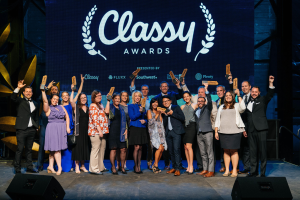 Gruppe von Menschen auf einer Bühne, die Auszeichnungen halten, mit Rednern darunter und einem Bildschirm im Hintergrund mit "Classy Awards 2019".