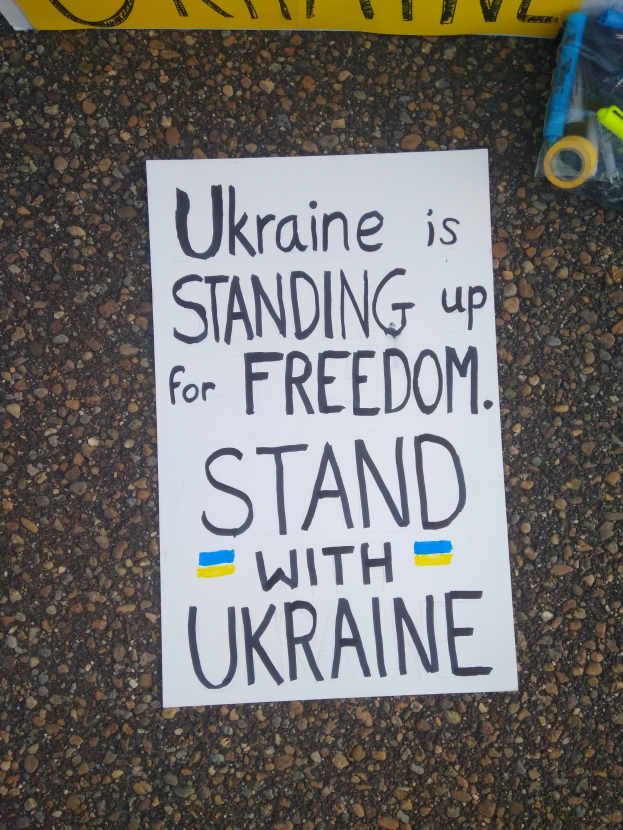 Weißer Papierbogen mit dem Text "Ukraine steht für Freiheit, steht mit Ukraine" auf dem Boden vor einem gelben Banner, mit einer Plastikhülle mit verschiedenen Gegenständen rechts.