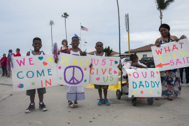 Eine Gruppe von Kindern hält Schilder und steht vor einem Gebäude, während eine Frau einen Kinderwagen schiebt; auf einem Schild steht: "Wir kommen in Frieden, gebt uns unseren Daddy."