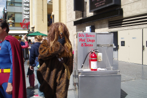 Eine Menschenmenge, darunter einige in Kostümen, versammelte sich vor einem Gebäude mit sichtbarer Beschilderung und Türen, mit einer Box und einem Feuerlöscher in der Nähe.
