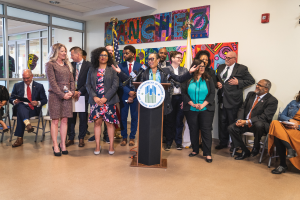 Eine Gruppe von Menschen, die in einem Raum mit einem Rednerpult, einem Mikrofon und einer Tafel mit der Aufschrift "Sancho-Stadtrat tritt dem Stadtrat bei" stehen und sitzen, mit Flaggen, einer Tafel, einem Feuerlöscher und Deckenlampen an der Rückwand.