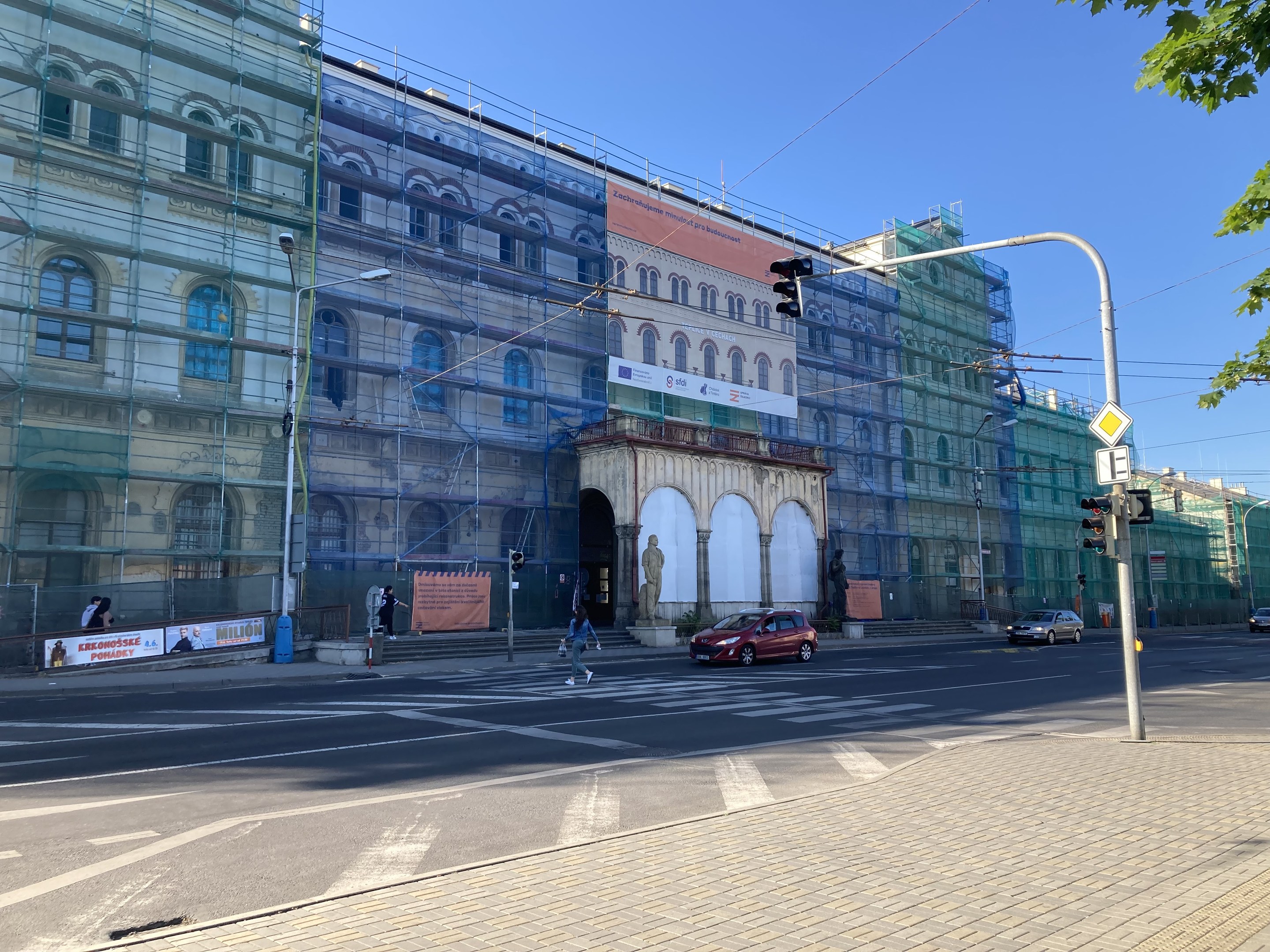 Großes Gebäude mit Gerüst während der Renovierung umgeben von Fahrzeugen, Fußgängern, Ampeln, Schildern, Straßeninfrastruktur, Bäumen und einem klaren blauen Himmel.