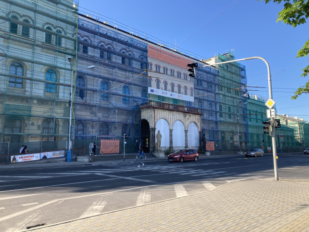 Großes Gebäude mit Gerüst während der Renovierung umgeben von Fahrzeugen, Fußgängern, Ampeln, Schildern, Straßeninfrastruktur, Bäumen und einem klaren blauen Himmel.