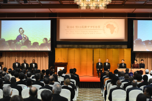 Eine Gruppe von Menschen, die in einem Raum mit einem Podium, Mikrofonen und einem großen Bildschirm sitzen, mit einer von einem Kronleuchter beleuchteten Decke darüber.