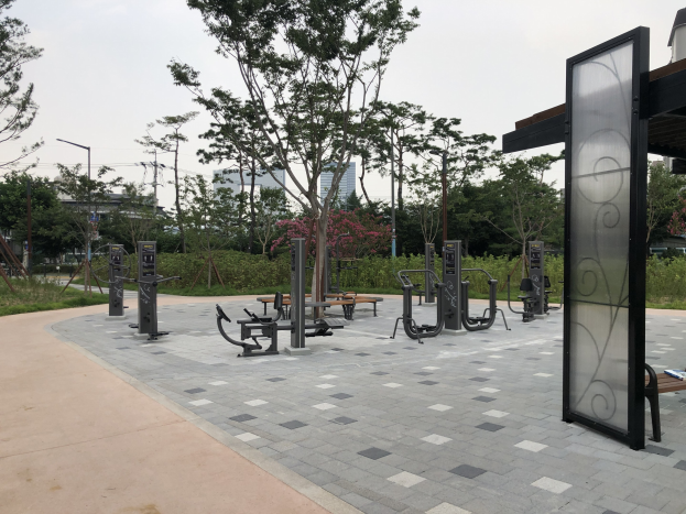Outdoor-Park mit Bänken, Bäumen, Pflanzen, Gras, Pfosten, Lichtern, Drähten und Gebäuden unter dem Himmel.
