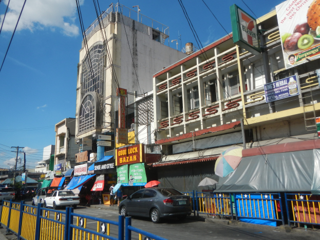 Eine belebte Stadtstraße mit Autos, hohen Gebäuden, Schildern, Regenschirmen, einem Metallzaun, Strommasten mit Drähten und einem bewölkten Himmel.