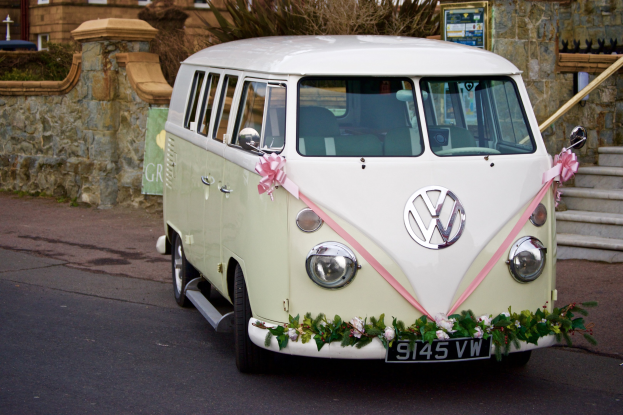 Ein weißer und pinker VW-Campervan mit floralen Verzierungen, der vor einer Steinmauer, einer Treppe und einem Gebäude geparkt ist.
