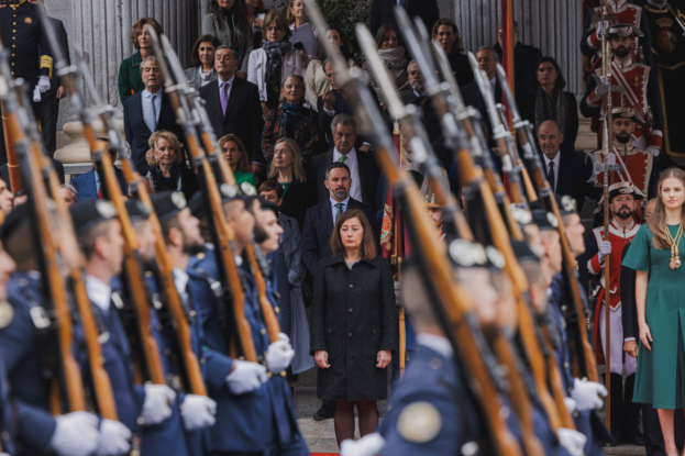 Eine Gruppe von Menschen in militärischer Kleidung steht vor einer Menge, einige halten Gewehre, bei einer Parade mit Säulen und einer Pflanze im Hintergrund.