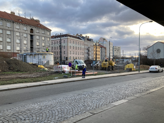 Stadtstraße mit parkenden Autos, Gebäuden, Laternen, Bäumen und einem Himmel mit Wolken, mit einer Sperre mit Fahrzeugen und Fussgängern im Vordergrund.