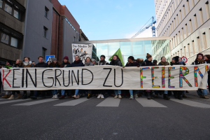 Eine Gruppe von Menschen steht auf einer Straße und hält ein Transparent mit der Aufschrift "Kein Grund zu Feiern" als Protest, im Hintergrund sind Gebäude, ein Schild, ein weiteres Transparent, ein Kran und ein bewölkter Himmel zu sehen.