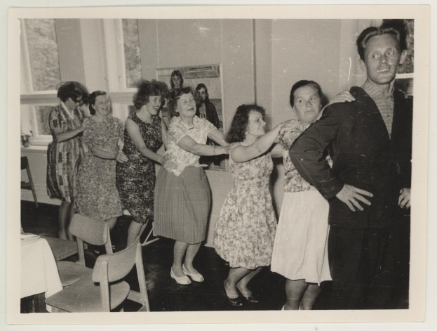 Eine Gruppe von Menschen, die in einem Raum im 1950er-Stil tanzen, mit Stühlen, einem Tisch mit einer weißen Decke, einem Foto Rahmen an der Wand und Fenstern im Hintergrund.