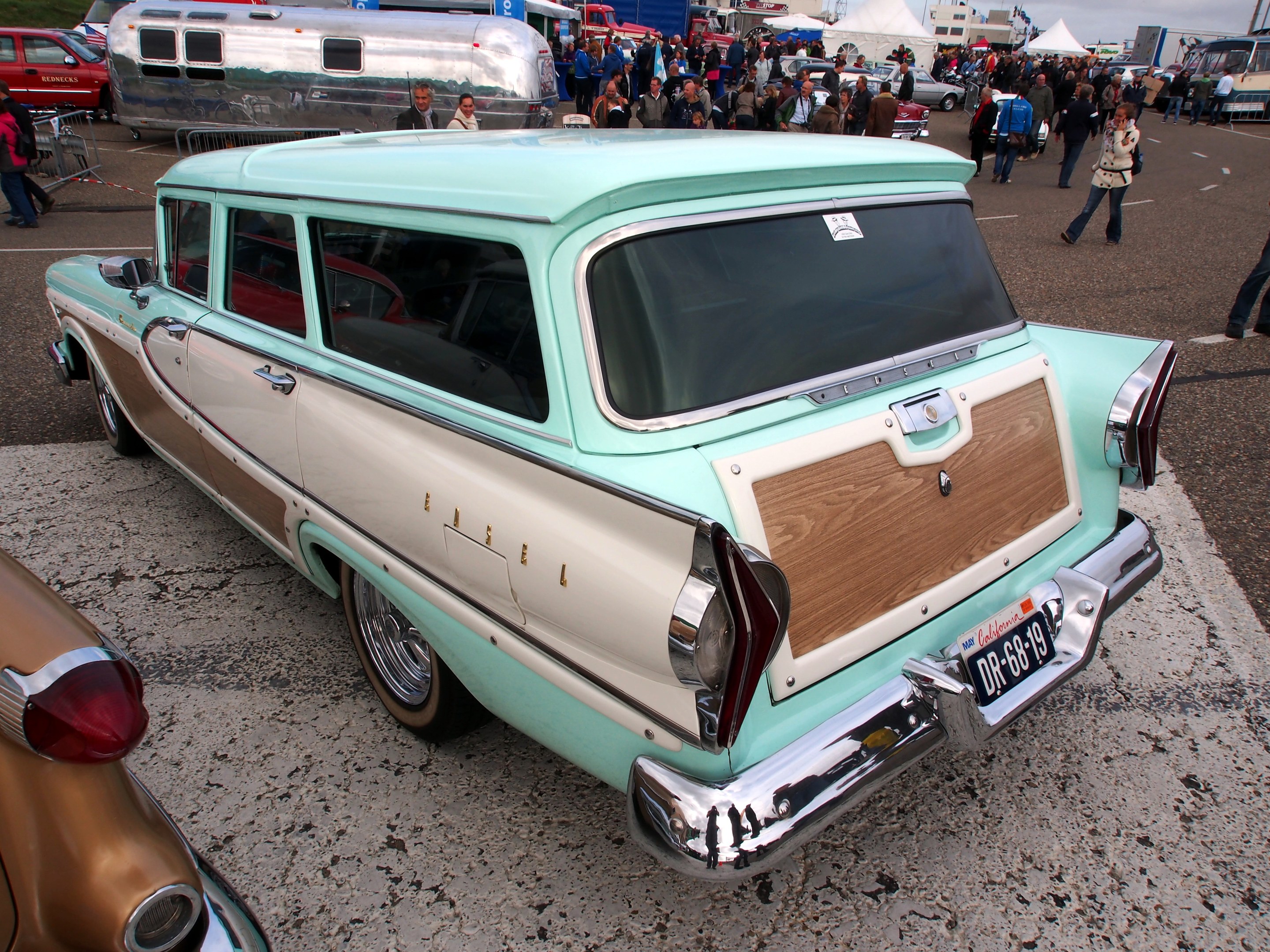 Ein grüner und weißer Kombiwagen auf einem belebten Parkplatz mit Zelten, Masten, Bäumen und einem klaren blauen Himmel im Hintergrund.