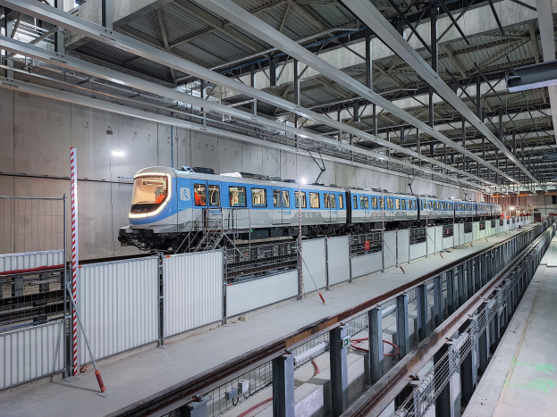 Ein blauer und weißer Zug, der durch eine große Indoor-Station mit einer Plattform auf der rechten Seite und einer Wand im Hintergrund fährt, unter einer Decke mit Stangen und Lampen.