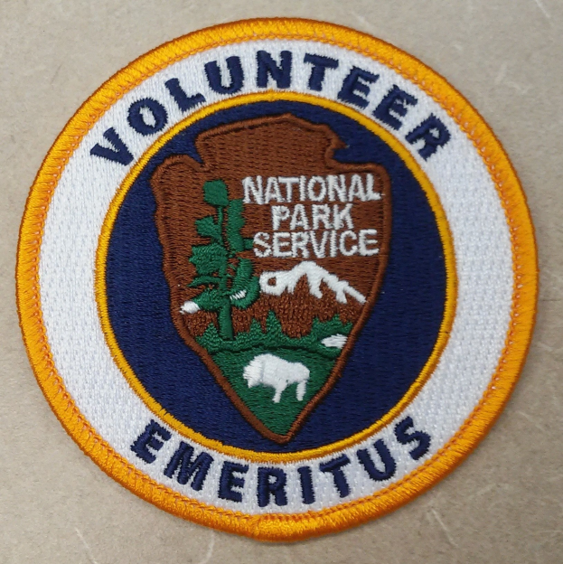 Runder Aufnäher mit "Volunteer National Park Service Emeritus" in schwarzer Schrift auf weißem Hintergrund, umrandet in blau mit einem gelben Kreis um den Text.
