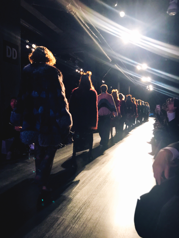 Eine Gruppe von Menschen läuft über den Laufsteg einer Fashion Show, während Zuschauer auf beiden Seiten zuschauen.