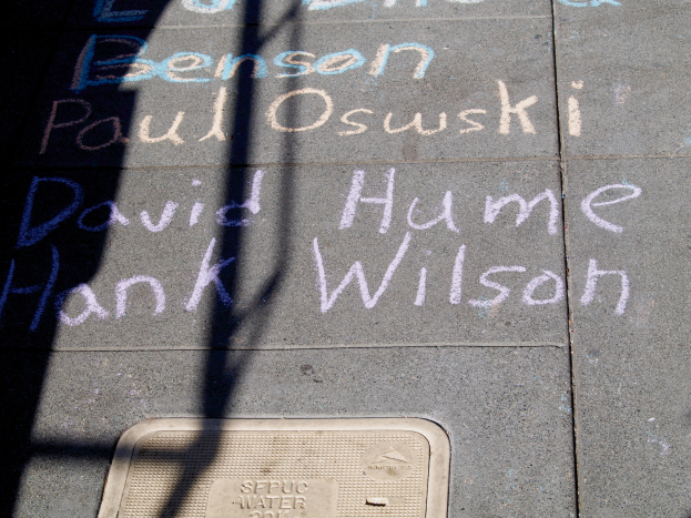 Ein Gehweg mit Kreide-Schrift, die wie ein Denkmal aussieht, eine Gulldeckel am Boden und ein Schatten eines Baums auf der linken Seite.