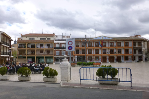 Ein belebter Stadtplatz mit Menschen, die sitzen und stehen, Topfpflanzen, einer Metallabsperrung, einem Schild an einem Pfahl, Straßenlaternen mit Fahnen, umliegenden Gebäuden mit Fenstern und einem bewölkten Himmel.