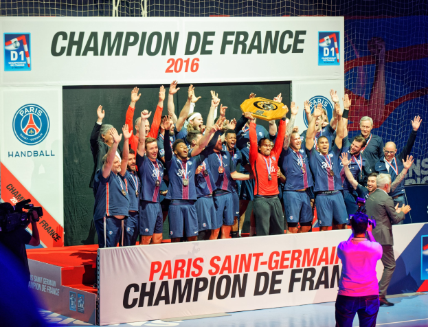 Eine Gruppe von Männern hält einen Pokal auf einer Bühne, während jemand filmt und ein "Champion de France 2016"-Schild im Hintergrund zu sehen ist.