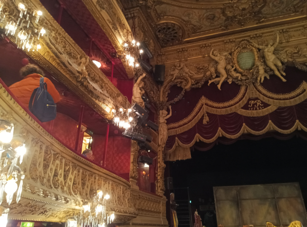Theaterinnenraum mit prunkvollen Kronleuchtern, Wandskulpturen und Menschen auf der Bühne, mit verstreuten Gegenständen, die eine lebendige Atmosphäre schaffen.