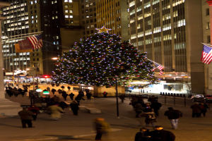 Ein Weihnachtsbaum auf einer Straße mit Menschen drum herum, flankiert von zwei Fahnenmasten, mit beleuchteten Gebäuden im Hintergrund.