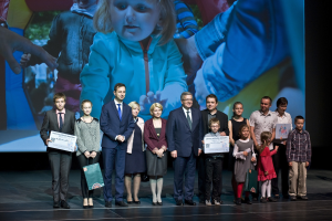 Gruppe von Menschen auf einer Buhne mit Taschen und Urkunden, im Hintergrund ein Bildschirm mit einem Kind und anderen