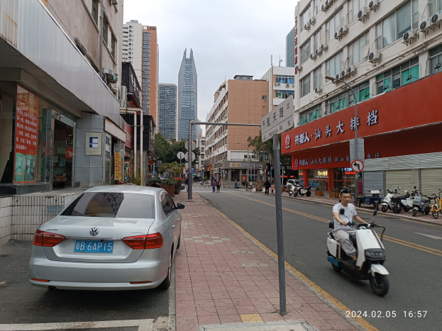 Ein Mann fährt mit einem Roller eine Straße mit hohen Gebäuden entlang, mit Menschen auf den Gehwegen und anderen Fahrzeugen auf der Straße, unter einem klaren blauen Himmel mit Bäumen im Hintergrund.