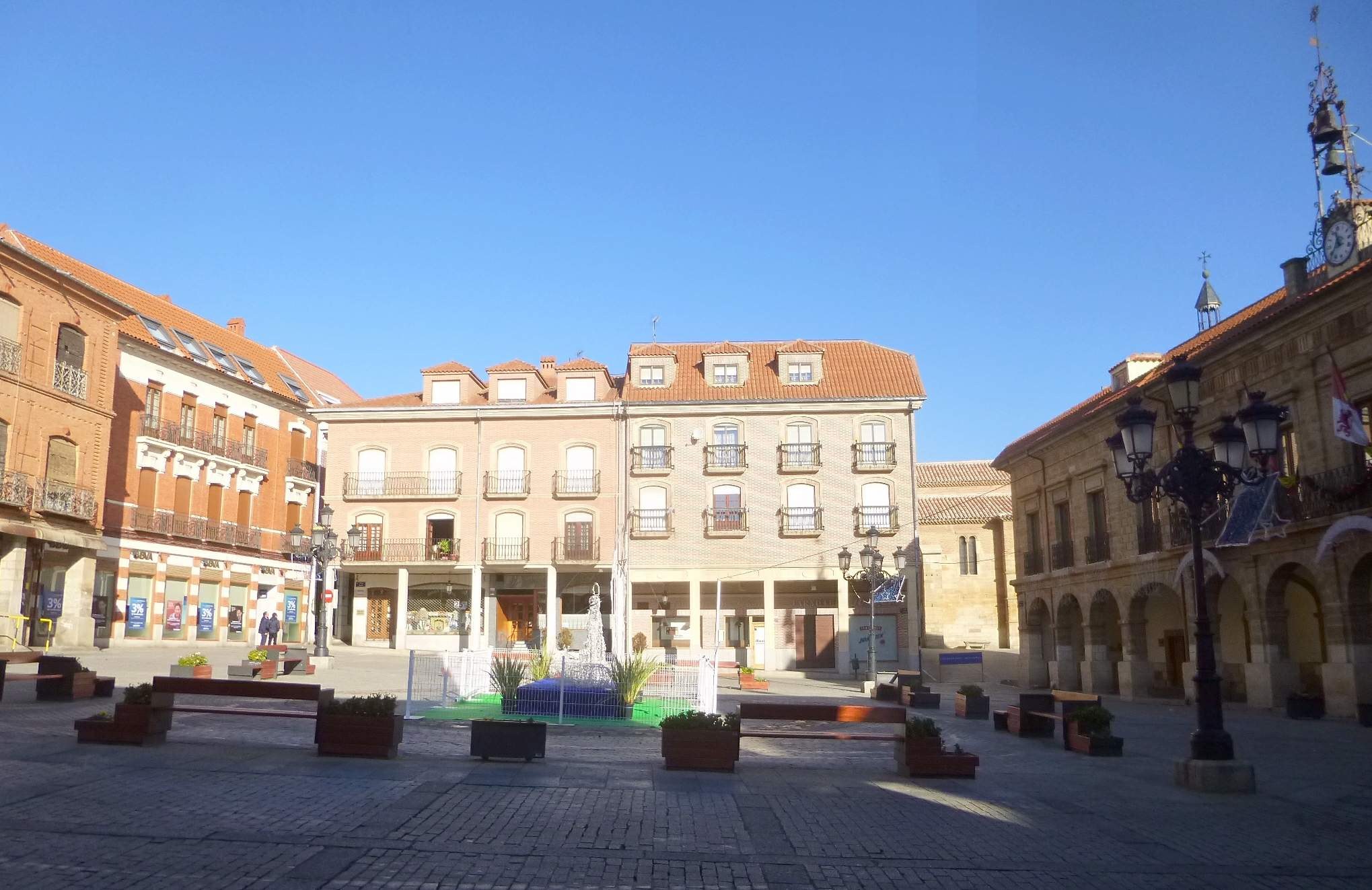 Ein Stadtplatz mit einem zentralen Brunnen, umgeben von Bänken, Topfpflanzen, Straßenlaternen und -masten, sowie einem Uhrenturm und Gebäuden im Hintergrund unter einem klaren blauen Himmel.