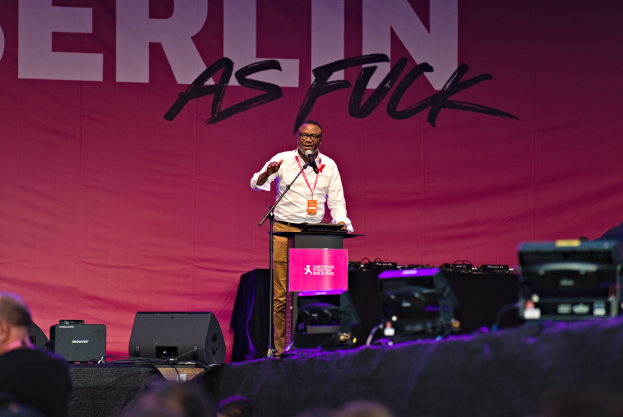 Mann in weißem Hemd und braunen Hosen hält Mikrofon auf Bühne, mit pinkem Banner 'Berlin as Fuck' im Hintergrund und Lautsprechern auf der Bühne.