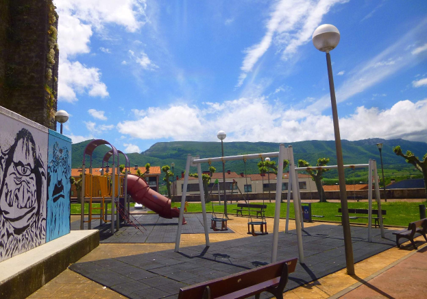 Ein Spielplatz mit Schaukeln, einer Rutsche, Bänken, Pfählen, Laternen, einer bemalten Wand, Gras, Bäumen, Gebäuden, Hügeln und einem bewölkten Himmel im Hintergrund.