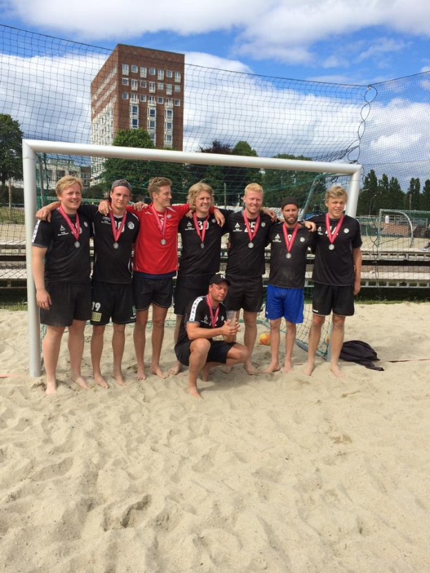 Eine Gruppe von Männern in Medaillen steht auf einem sandigen Strand, umgeben von einem Netz, mit Bäumen, Gebäuden und einem bewölkten Himmel im Hintergrund.