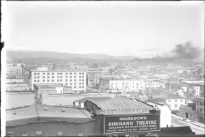 Ein Schwarz-Weiß-Bild eines Fabrikgebäudes mit Rauch, der aus seinem Schornstein aufsteigt und von Gebäuden umgeben ist.
