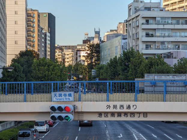 Eine Straße mit fahrenden Autos, eine Brücke mit Geländern und ein Schild, Verkehrsampeln, Laternen, Bäume und Gebäude mit Fenstern unter einem klaren Himmel.