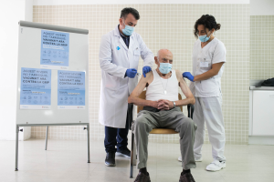 Ein älterer Mann sitzt in einem Krankenhauszimmer, während ihm zwei maskierte und handschuhgetragene Ärzte ein Vakzin verabreichen, mit einer Tafel links und Gegenständen auf einer Plattform rechts.