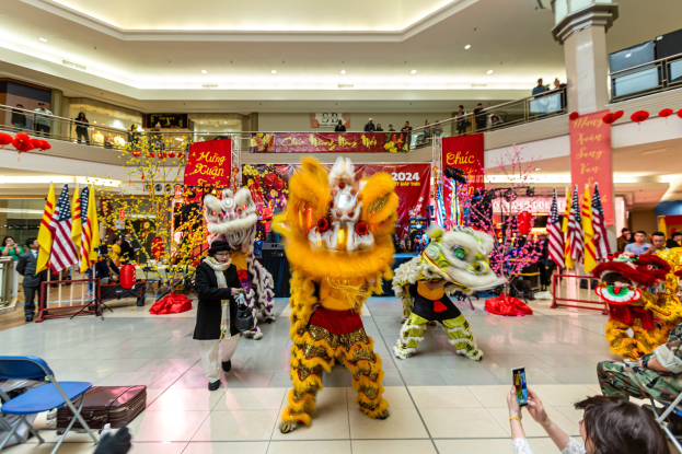 Eine lebendige chinesische Neujahrsfeier in einem Einkaufszentrum mit Menschen in Kostümen, die tanzen, und anderen, die auf Stühlen sitzen und mobile Telefone halten, umgeben von Flaggen, Bannern, Dekorationen und Lichtern.