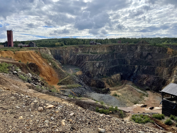Großer Tagebau auf einem Schotterfeld umgeben von Bäumen, Gebäuden und einem Schuppen, mit einem bewölkten Himmel und einem mit Steinen und Pflanzen bedeckten Boden.