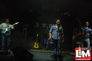 Drei Personen auf der Bühne, eine spielt Gitarre und eine singt in ein Mikrofon, mit einem Snaredrum-Arrangement und Beleuchtung im Hintergrund.