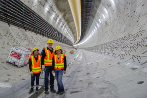 Drei Personen in Sicherheitsoveralls und gelben Helmen stehen in einem Tunnel, mit einem weißen Kasten links und Rohren rechts.