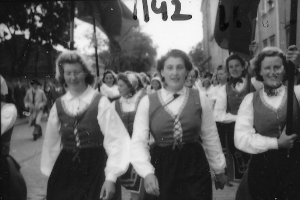 Eine Gruppe von Frauen in traditioneller bayerischer Tracht spaziert strahlend durch eine Straße, umgeben von Bäumen und Gebäuden, auf einem Schwarz-Weiß-Bild.