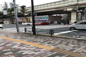 Eine Stadtstraße mit Fahrzeugen, Fußgängern an einer Ampel, einem Gehweggeländer, Verkehrszeichen, einer Brücke, Bäumen, Gebäuden und einem klaren blauen Himmel.