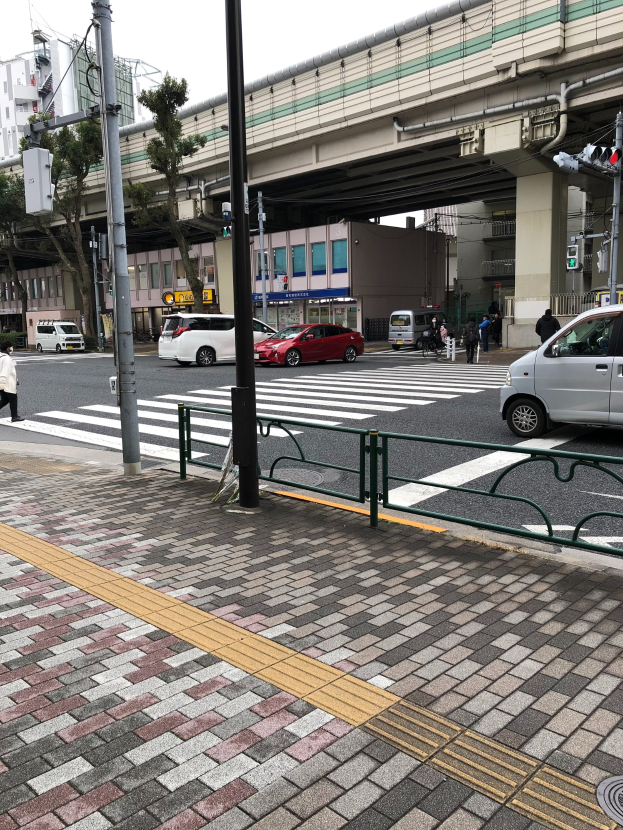 Eine Stadtstraße mit Fahrzeugen, Fußgängern an einer Ampel, einem Gehweggeländer, Verkehrszeichen, einer Brücke, Bäumen, Gebäuden und einem klaren blauen Himmel.
