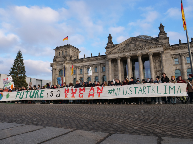 Gruppe von Menschen vor dem Reichstagsgebäude in Berlin mit einer Fahne mit der Aufschrift "Zukunft ist ein Menschenrecht" und umgeben von Bäumen und Fahnenmasten.