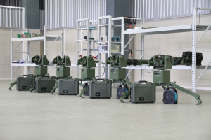 Eine Gruppe von Militärrobotern ist auf einem Boden positioniert, umgeben von Koffern und anderen Gegenständen auf Gestellen, mit einer Wand und einem Geländer im Hintergrund.