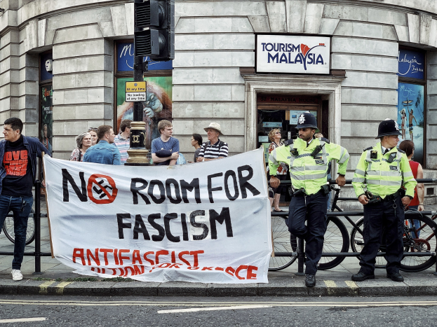 Eine Gruppe von Menschen mit einer 'Kein Platz für Faschismus'-Plakette, vor der zwei Polizeibeamte auf einem Gehweg stehen, mit Fahrrädern, einer Ampel und einem Gebäude im Hintergrund.