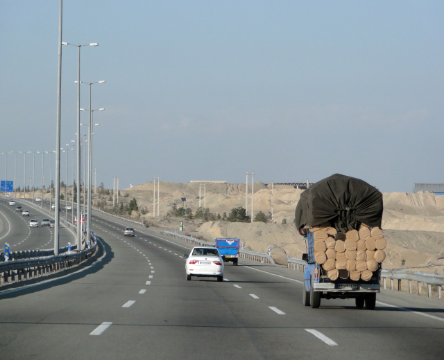 Ein Holzlaster mit einer großen Ladung Holz fährt auf einer Autobahn mit Geländern, Laternen, Schildern, Bäumen und Sand, mit Hügeln und einem klaren blauen Himmel im Hintergrund.