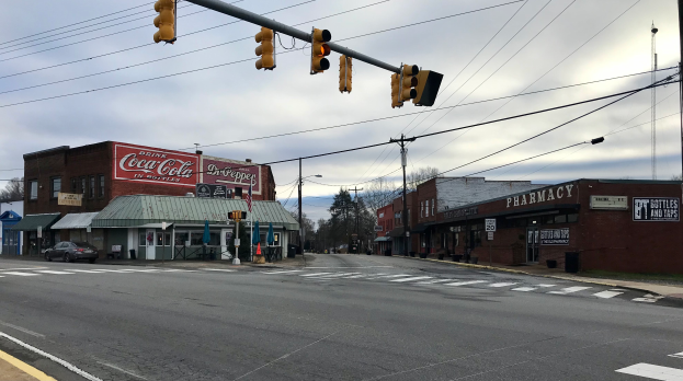 Eine Stadtstra├čenkreuzung mit Ampel, Fahrzeugen, Geb├Ąuden mit Namensschildern, Strommasten mit Kabeln, B├Ąumen und einem bew├Âlktem Himmel.