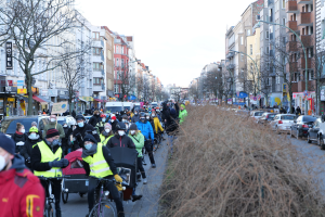 Eine große Gruppe von Menschen mit Masken und Sicherheitswesten fährt auf Fahrrädern eine von Bäumen gesäumte Straße mit Gebäuden, Laternenmästen und Texttafeln entlang, während Fahrzeuge die Straße teilen und trockenes Gras die rechte Seite unter einem klaren blauen Himmel einnimmt.