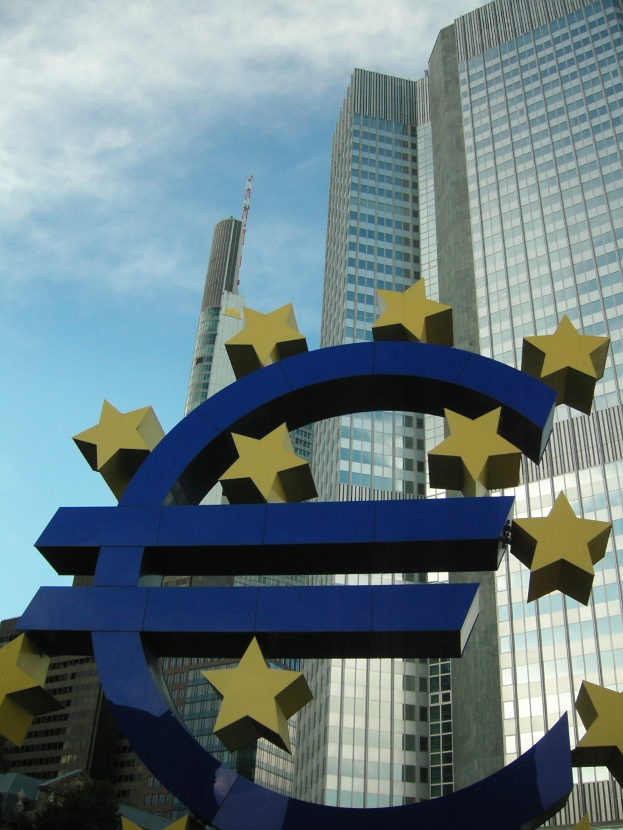 Euro-Zeichen vor dem Gebäude der Europäischen Zentralbank in Frankfurt, Deutschland, mit Bäumen und einem bewölkten Himmel.